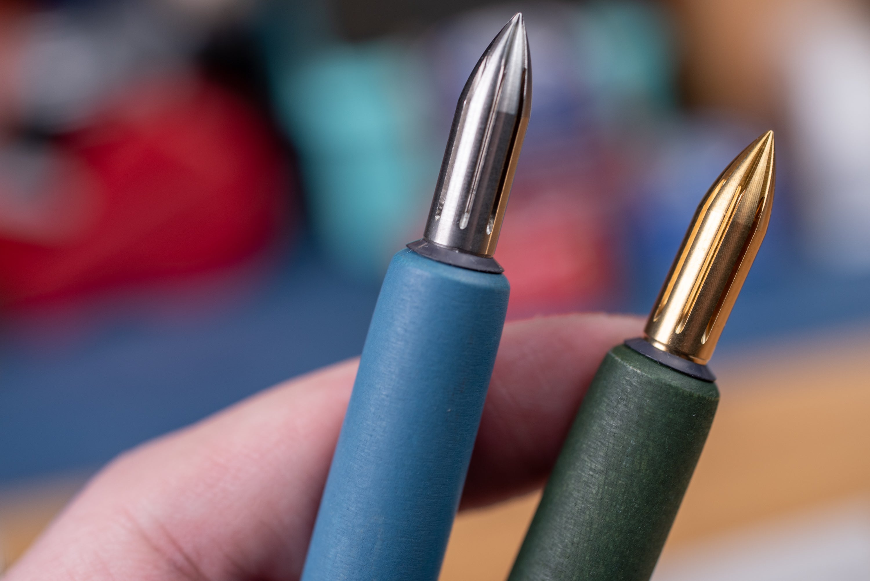 Two pen nibs, one blue and one green, held in a hand with a blurred background.